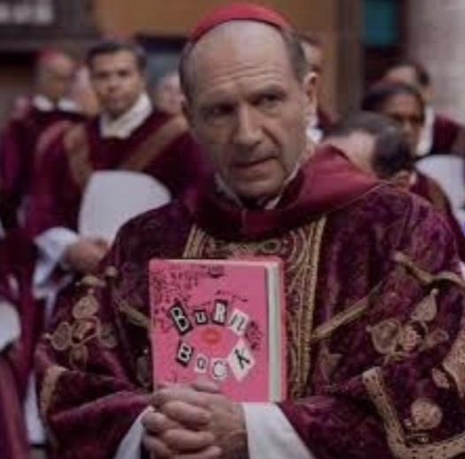 Na imagem, um plano médio de um homem de meia-idade (o ator Ralph Fiennes), caracterizado como um Cardeal ou autoridade religiosa, vestindo vestes luxuosas de veludo vinho ou roxo e segurando um objeto rosa choque. O objeto é o 