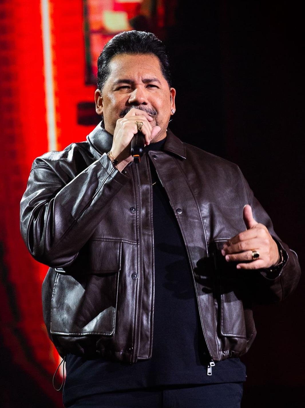 Homem cantando no palco com microfone na mão, usando jaqueta de couro preta, em apresentação musical ao vivo.