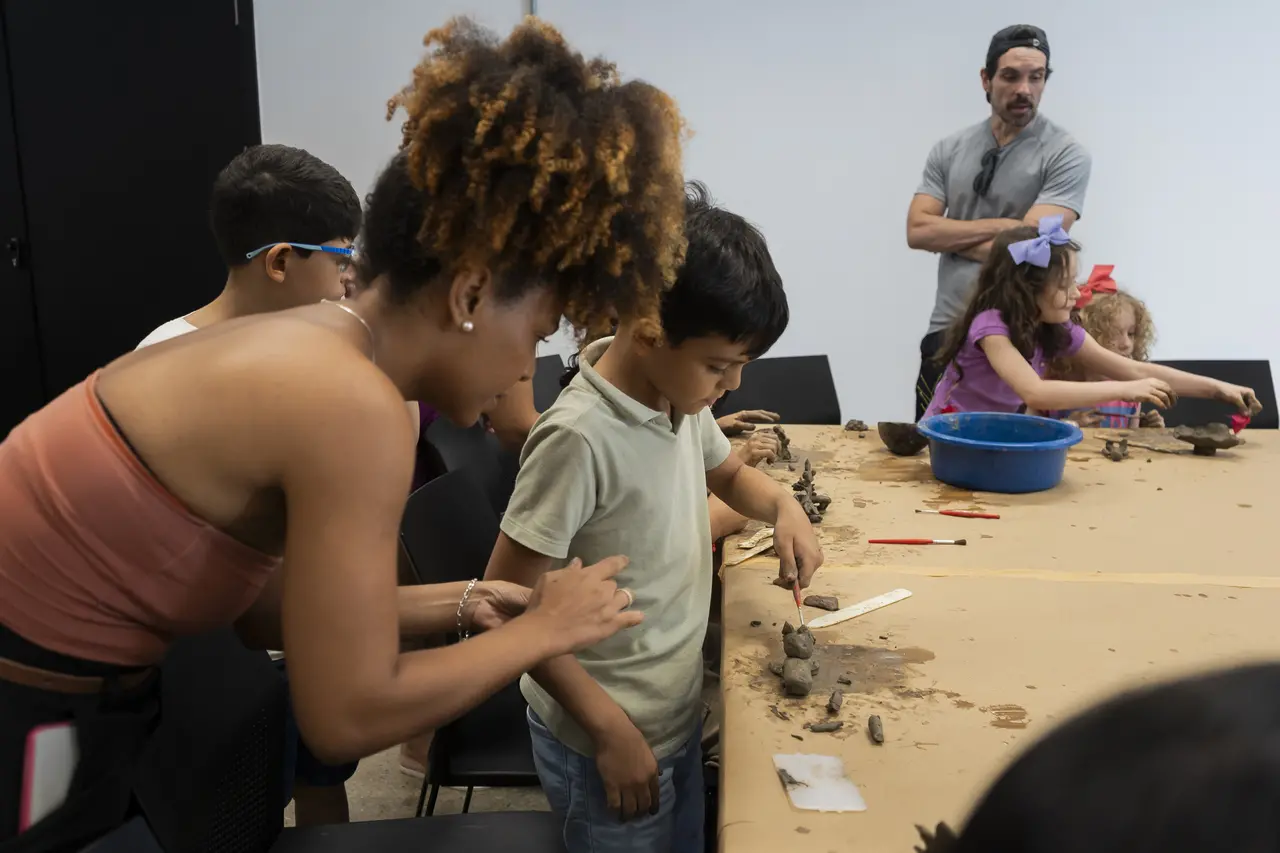 Uma arte-educadora ajuda um menino em uma mesa com argila e ferramentas, enquanto outras crianças e um adulto observam ao redor.