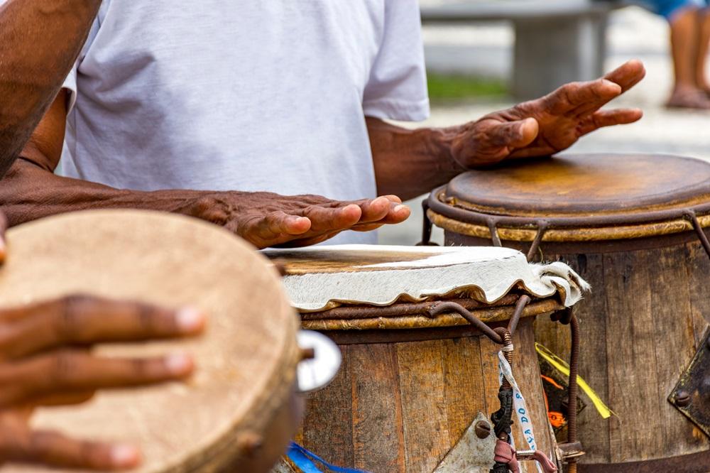 Na imagem, mãos de um músico tocando dois atabaques de madeira durante uma roda de Capoeira ou apresentação cultural. A pele de um dos tambores é de cor clara, enquanto a outra é mais escura. O músico veste uma camiseta branca simples.