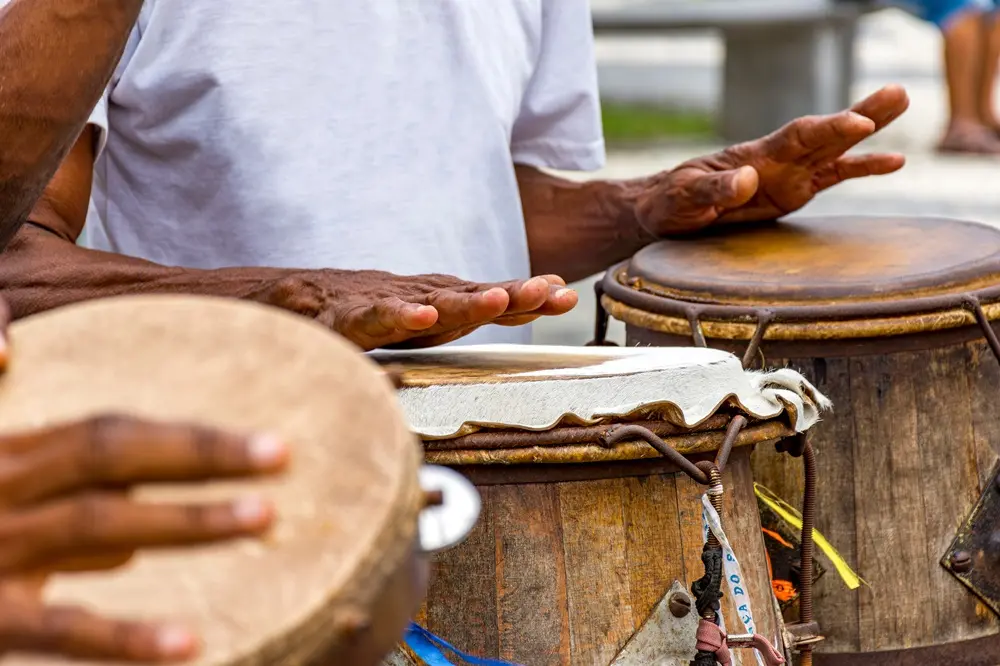 Na imagem, mãos de um músico tocando dois atabaques de madeira durante uma roda de Capoeira ou apresentação cultural. A pele de um dos tambores é de cor clara, enquanto a outra é mais escura. O músico veste uma camiseta branca simples.