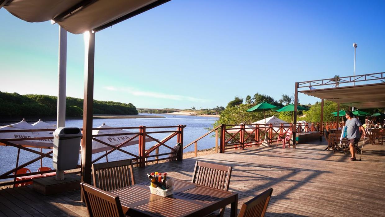 Na imagem, vista ensolarada de um restaurante à beira-rio ou beira-mar, com um deck de madeira marrom claro. Mesas e cadeiras de madeira escura estão dispostas sob uma área de cobertura. No primeiro plano, uma mesa para quatro pessoas é vista. O deck é separado da água por uma cerca de madeira. Em frente ao deck, há uma ampla faixa de água calma que se encontra com a vegetação verdejante em ambas as margens. Uma ponte pode ser vista à distância sobre o rio, sob um céu azul claro. Um homem com uma camisa azul clara e shorts está em pé na parte direita do deck.
