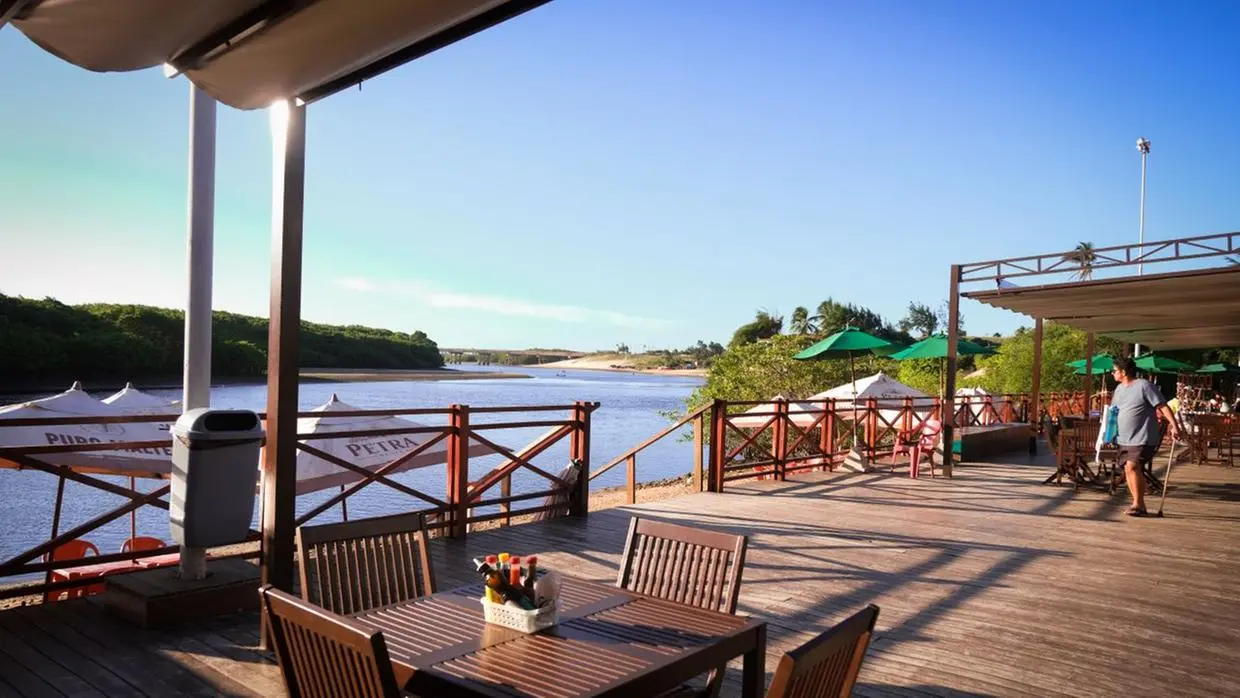 Na imagem, vista ensolarada de um restaurante à beira-rio ou beira-mar, com um deck de madeira marrom claro. Mesas e cadeiras de madeira escura estão dispostas sob uma área de cobertura. No primeiro plano, uma mesa para quatro pessoas é vista. O deck é separado da água por uma cerca de madeira. Em frente ao deck, há uma ampla faixa de água calma que se encontra com a vegetação verdejante em ambas as margens. Uma ponte pode ser vista à distância sobre o rio, sob um céu azul claro. Um homem com uma camisa azul clara e shorts está em pé na parte direita do deck.