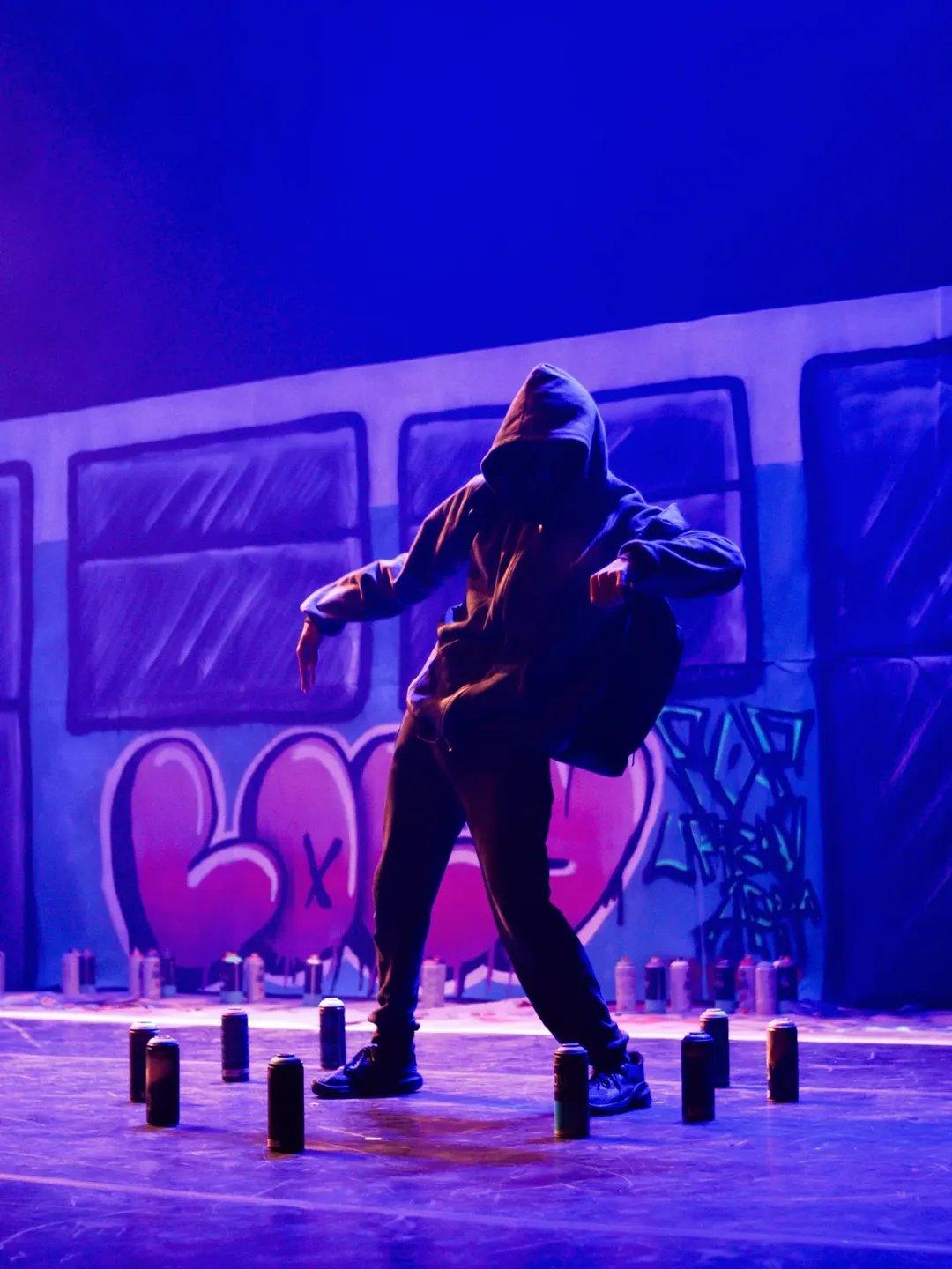 Na imagem, um artista de dança de rua, com o rosto escondido pelo capuz, é iluminado por luzes azuis e roxas em um palco. O dançarino está em uma postura dinâmica, curvado, com as pernas afastadas e os braços estendidos, vestido com roupas escuras largas. O cenário é um mural de graffiti estilizado, que se assemelha à lateral de um trem, com janelas escuras e a palavra 