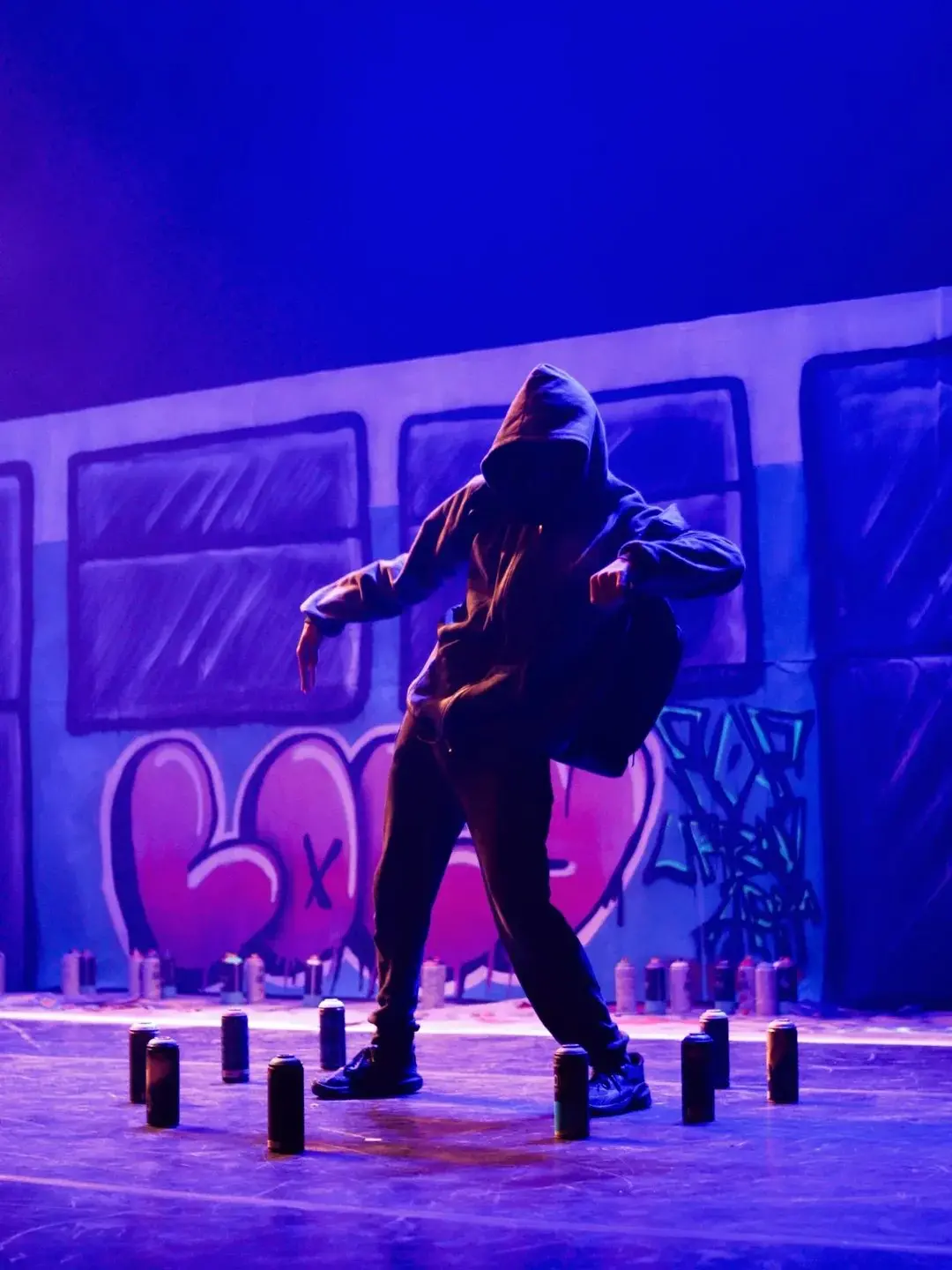 Na imagem, um artista de dança de rua, com o rosto escondido pelo capuz, é iluminado por luzes azuis e roxas em um palco. O dançarino está em uma postura dinâmica, curvado, com as pernas afastadas e os braços estendidos, vestido com roupas escuras largas. O cenário é um mural de graffiti estilizado, que se assemelha à lateral de um trem, com janelas escuras e a palavra 