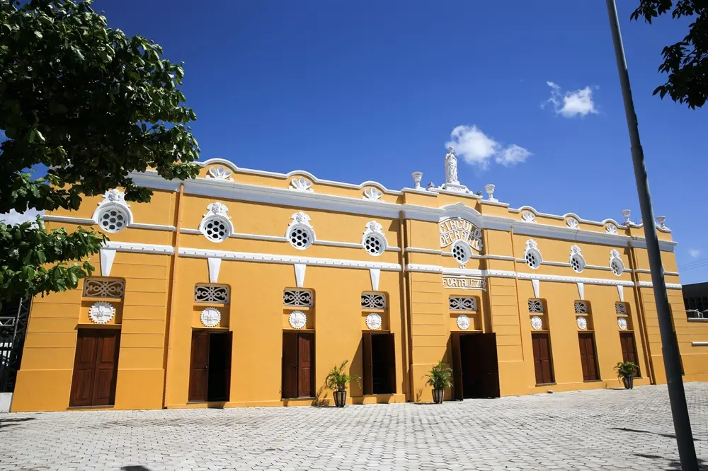 Na imagem, vista externa em ângulo aberto e sob a luz do dia do histórico Teatro São José em Fortaleza. O edifício é característico, pintado em uma cor amarela viva, com detalhes arquitetônicos e caixilhos de janelas brancos. A fachada apresenta uma série de portas de madeira escura e janelas redondas em estilo olho de boi. A platibanda superior é decorada com relevos em massa branca e uma estátua branca de Nossa Senhora ou uma figura clássica. O centro do edifício possui uma placa redonda no topo das portas centrais com o nome do teatro. O chão em frente ao teatro é pavimentado com blocos de concreto branco. À esquerda e à direita superior, há folhagem verde de árvores, e o céu é de um azul profundo e claro com poucas nuvens.