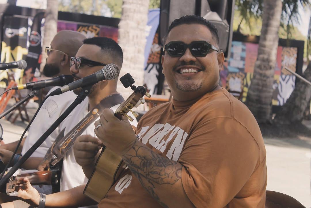 Na imagem, uma fotografia externa e bem iluminada capta um músico em primeiro plano tocando um cavaquinho e sorrindo. O homem, que tem barba e bigode ralos, veste uma camiseta cor de tijolo com estampas brancas e óculos de sol escuros. Ele tem tatuagens visíveis no braço direito, que segura o instrumento. À sua esquerda, fora de foco, outros dois músicos, ambos de óculos de sol, tocam violões. Um microfone está posicionado em frente a eles. O fundo sugere um ambiente de show ao ar livre, com troncos de palmeiras, luz do sol filtrada e o que parece ser um banner colorido ou arte.