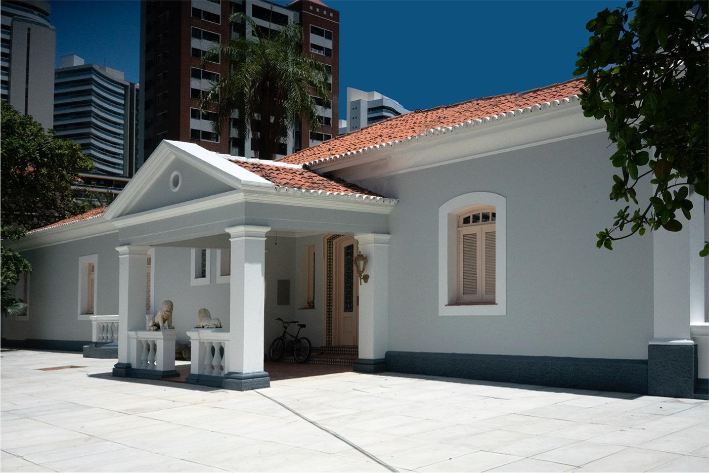 Na imagem, uma fotografia da fachada de um prédio histórico de cor cinza-clara, que abriga o Museu da Imagem e do Som (MIS) do Ceará. O edifício tem um telhado de telhas de barro alaranjadas e uma varanda de entrada sustentada por colunas brancas, com um frontão triangular. Dois pequenos ornamentos de leão de pedra estão sobre os parapeitos laterais da varanda. Uma bicicleta está estacionada perto da porta de entrada. O piso externo é de concreto claro e liso. No fundo, elevando-se sobre o prédio baixo e tradicional, está a paisagem urbana de Fortaleza, com vários edifícios modernos e altos de vidro e concreto. O céu é azul claro.