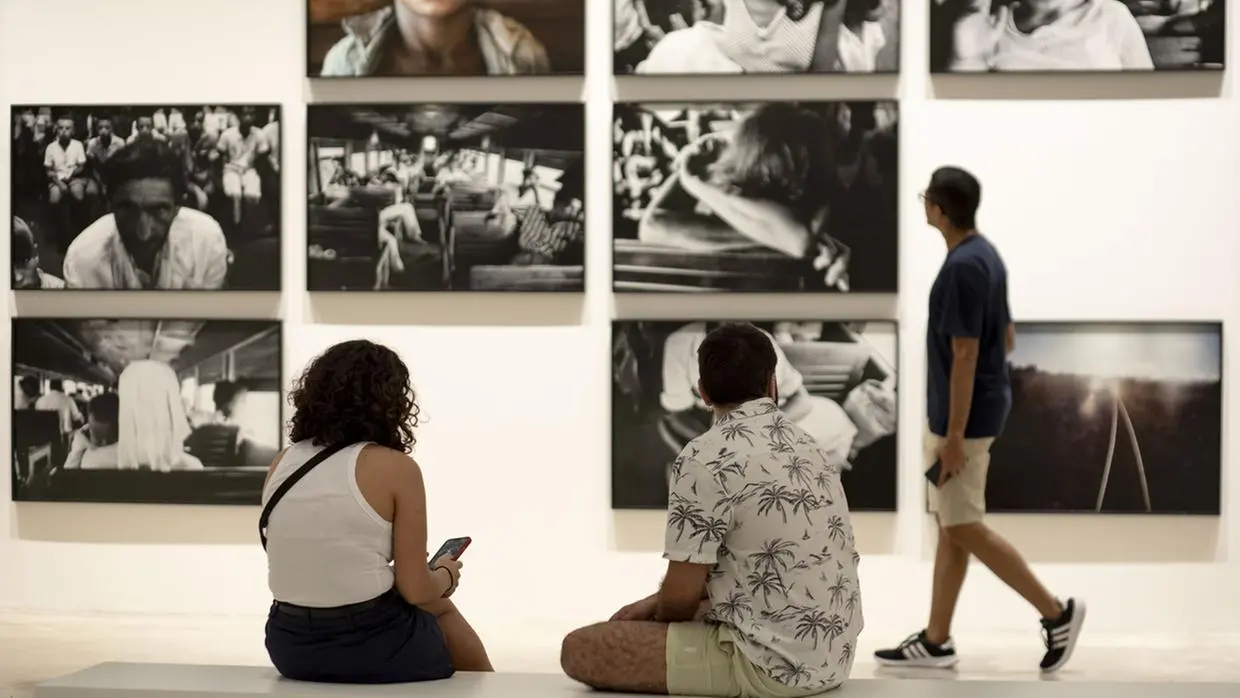 Na imagem, vista interna de uma galeria de arte ou museu, provavelmente a Pinacoteca do Ceará, onde várias fotografias estão penduradas em uma parede branca. As fotografias estão emolduradas em preto e a maioria é em preto e branco com alto contraste, retratando pessoas e cenas. No primeiro plano, à esquerda, um homem e uma mulher estão sentados em um banco, de costas para o observador, olhando para o celular. A mulher veste uma regata branca e shorts escuros; o homem veste uma camisa estampada com folhas de palmeira em fundo branco e shorts claros. À direita, um terceiro visitante, vestindo uma camiseta azul escura e shorts claros, está caminhando ao longo da parede, olhando para as obras.