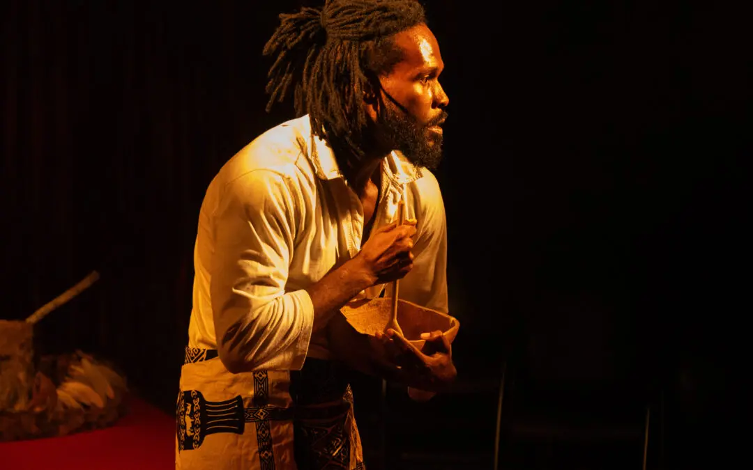 Na imagem, um close-up de um ator em um palco escuro, iluminado por uma luz intensa e quente que destaca sua figura. O ator, um homem negro com longos dreadlocks amarrados e barba cheia, está em perfil, olhando para fora da cena com uma expressão concentrada. Ele veste uma camisa branca de mangas compridas e calças ou um tecido envolvente de cor clara com um padrão geométrico escuro. Ele segura uma tigela ou cuia de madeira e um utensílio, como se estivesse executando um ritual ou preparando algo. O fundo é predominantemente preto, com uma cortina escura visível atrás e um tapete ou tecido vermelho no chão na parte inferior esquerda.