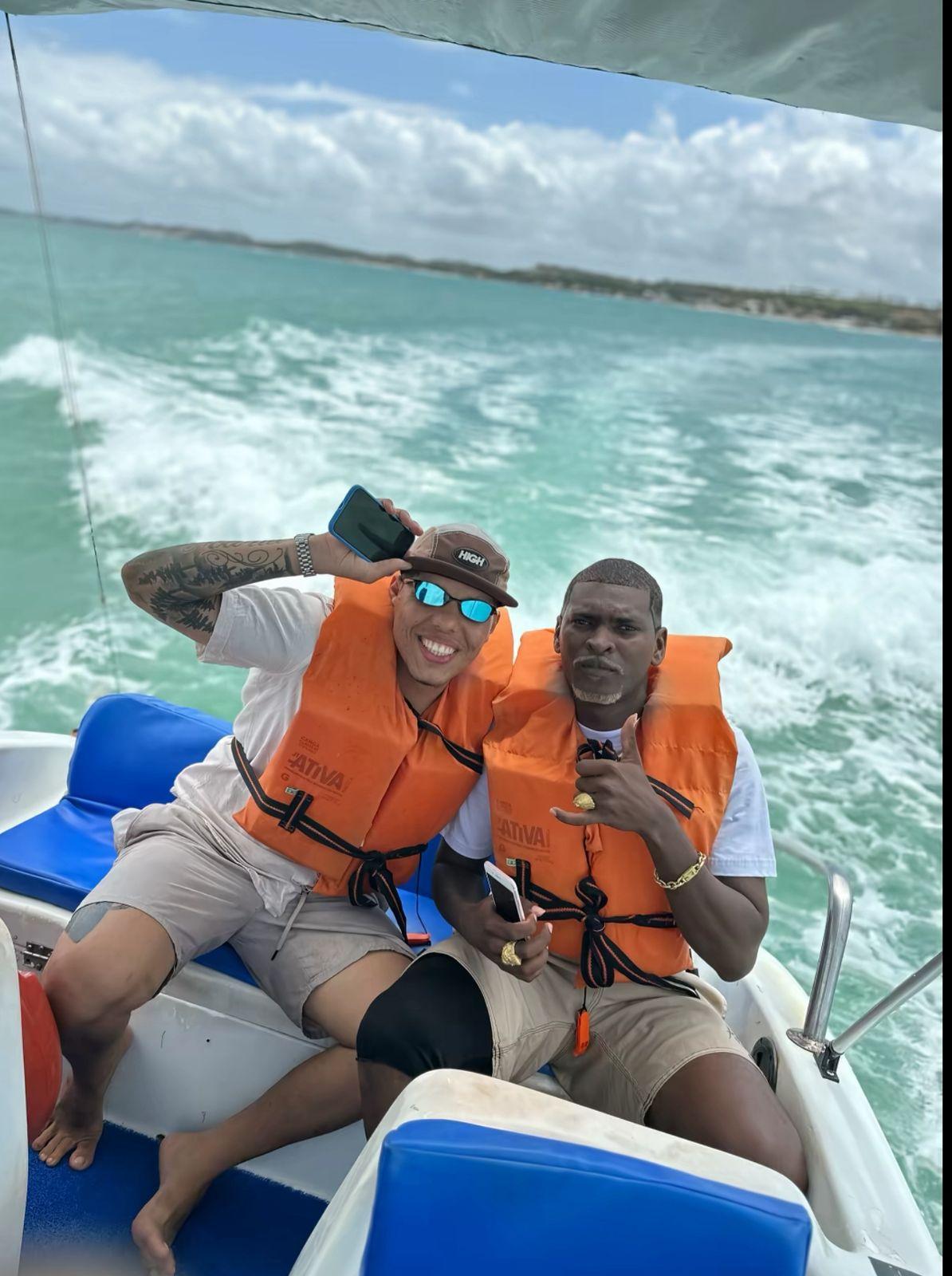 Dois homens em um passeio de lancha no mar, usando coletes salva-vidas laranja, sorrindo e fazendo gestos de diversão, com a água e o céu ao fundo.