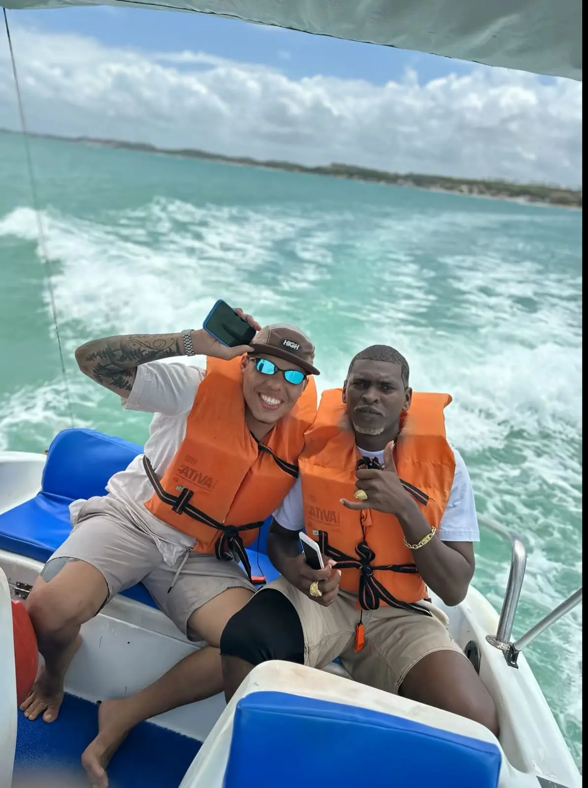 Dois homens em um passeio de lancha no mar, usando coletes salva-vidas laranja, sorrindo e fazendo gestos de diversão, com a água e o céu ao fundo.