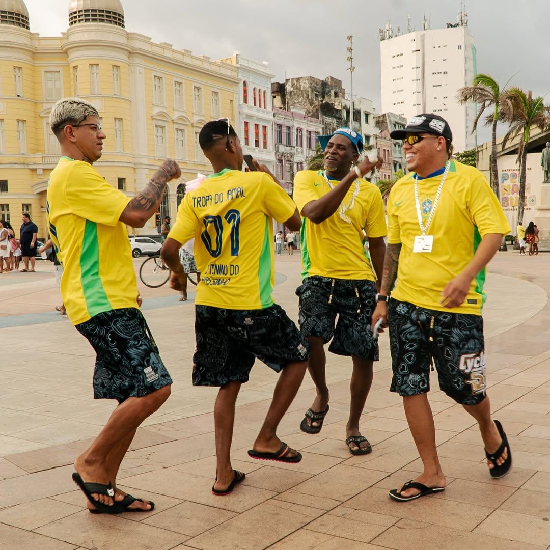 Grupo de amigos comemorando e dançando na rua durante evento no Recife, usando camisetas amarelas e shorts com estampas, com prédios históricos ao fundo.