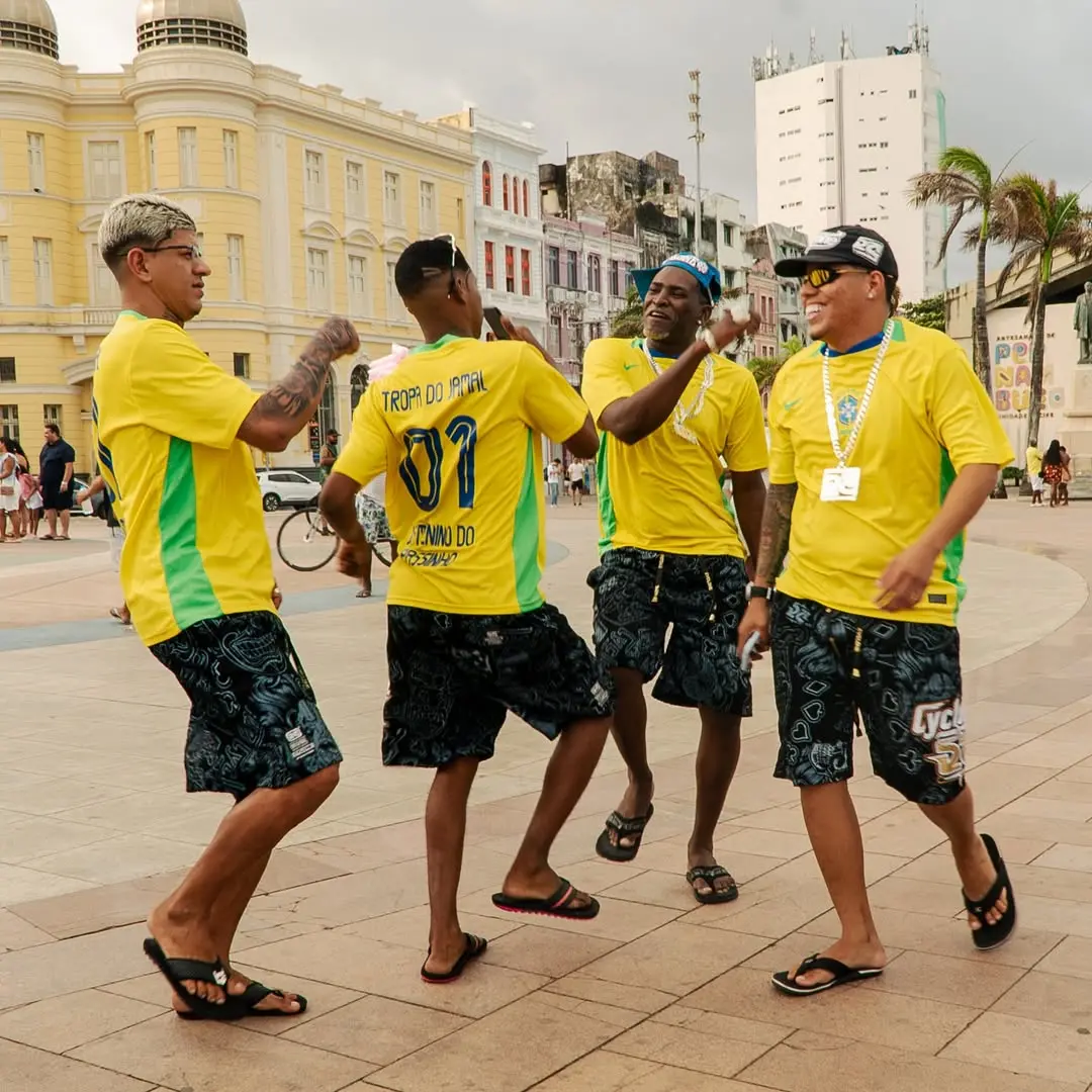 Grupo de amigos comemorando e dançando na rua durante evento no Recife, usando camisetas amarelas e shorts com estampas, com prédios históricos ao fundo.