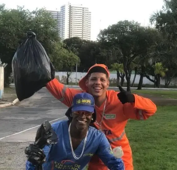 Dois catadores de lixoapós concluírem uma corrida beneficente, carregando sacos de lixo reciclável, em um parque urbano com árvores e prédios ao fundo.