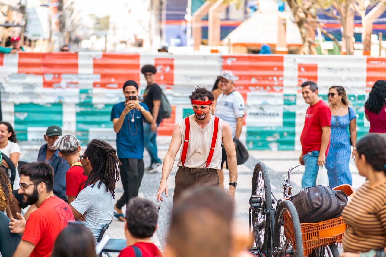 Um artista de palhaçaria, sorri usando suspensórios vermelhos e uma bandana vermelha, atuando para uma plateia reunida em uma área externa no centro de uma cidade. Há pessoas ao fundo, algumas caminhando e outras observando.