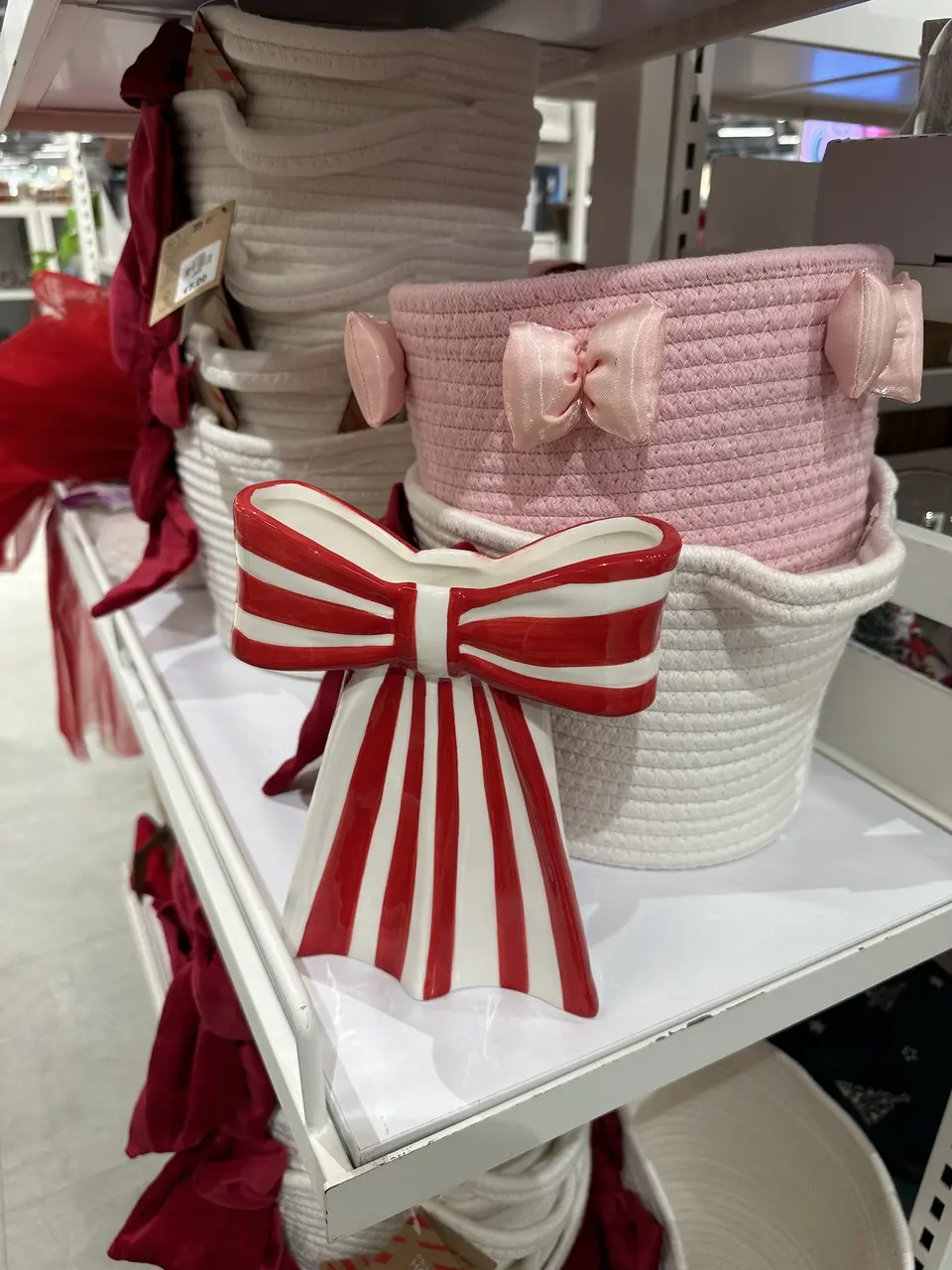 Uma imagem de prateleiras de loja exibindo artigos de decoração. Em primeiro plano, um objeto decorativo em formato de laço grande nas cores branco e vermelho com listras verticais e horizontais, apoiado em uma prateleira branca. Ao fundo, há pilhas de cestos de armazenamento feitos de corda trançada; um cesto é rosa-claro com laços de tecido rosa nas laterais, e os demais são brancos. Peças de tecido vermelho, possivelmente laços ou fitas, aparecem na lateral esquerda, reforçando o tema de decoração.