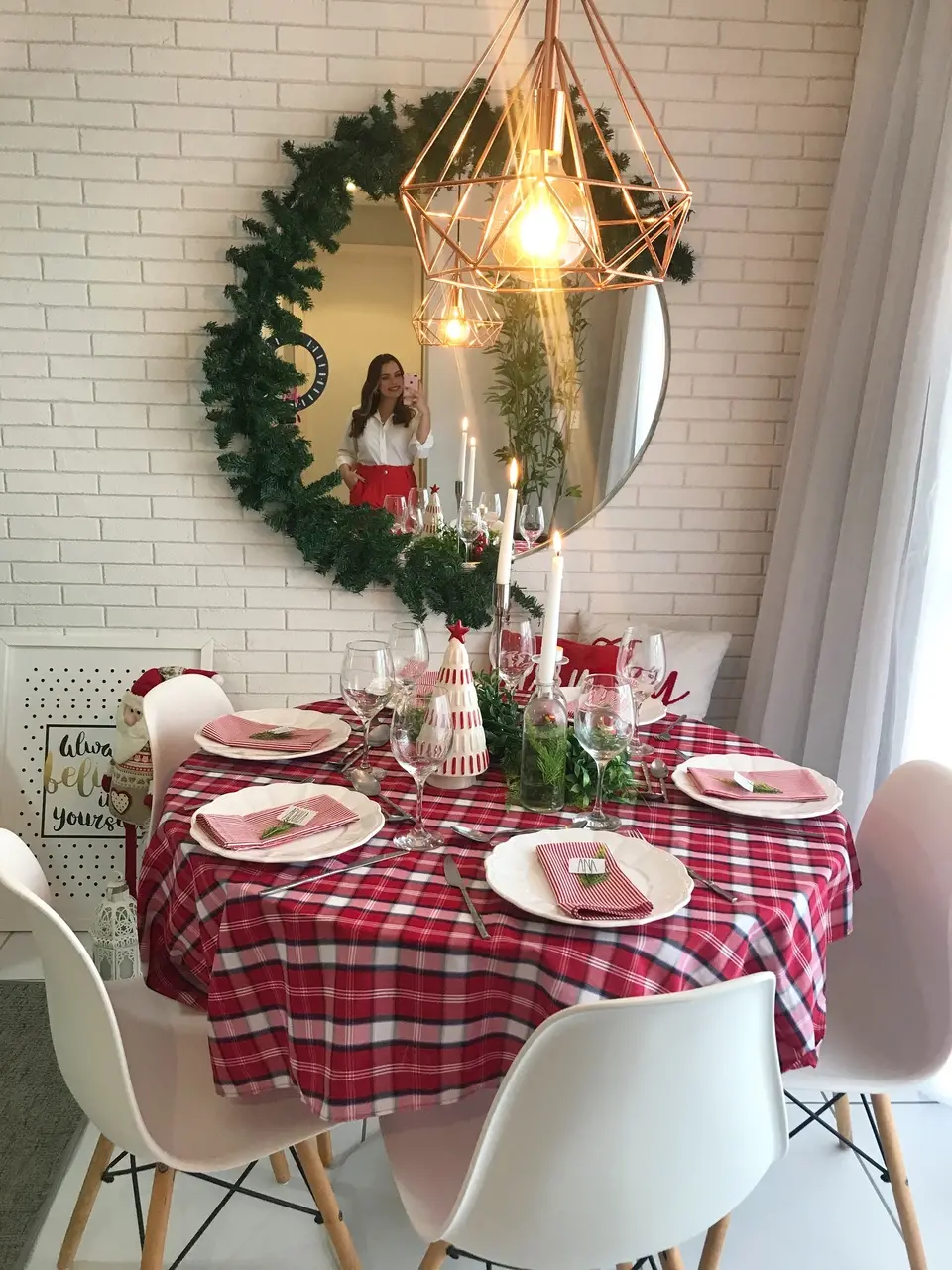 Uma mesa de jantar redonda decorada com o tema Natalino. A mesa é coberta por uma toalha xadrez vermelha e branca. Ela está posta com pratos brancos e guardanapos xadrez (vermelho e branco) com detalhes de folhagem. O centro da mesa possui velas brancas acesas em diferentes alturas e um adorno em formato de árvore listrada. Acima da mesa, há um lustre moderno com estrutura geométrica em tom de cobre. Ao fundo, uma parede de tijolos brancos apresenta um espelho redondo grande, emoldurado por uma guirlanda de folhagens verdes. O reflexo no espelho mostra a colunista Ana Karenyna, uma mulher vestida de branco e vermelho tirando a foto.