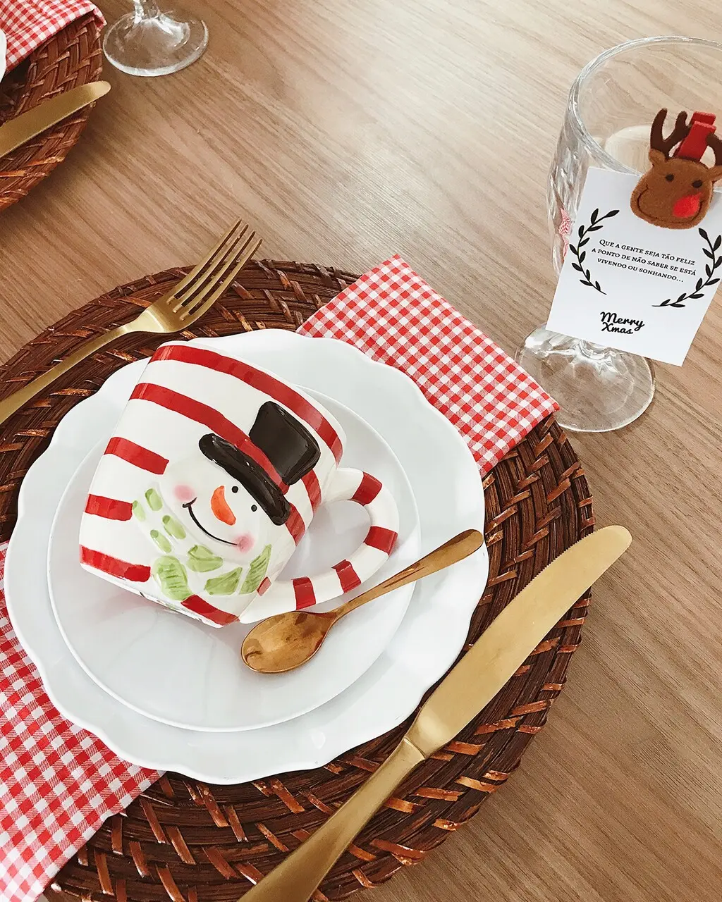 Uma foto em close de um lugar à mesa com o tema Natal. Sobre uma mesa de madeira clara, há um sousplat de fibra trançada marrom escura. O sousplat apoia dois pratos brancos, e sobre eles está uma caneca em formato de boneco de neve, listrada em vermelho e branco, com o rosto do boneco e um chapéu preto pintados. Os talheres são dourados, sendo uma colher de madeira pequena e os talheres principais dispostos nas laterais. Um guardanapo xadrez vermelho e branco está dobrado sob os pratos. À direita, uma taça de vinho possui um tag de presente pendurado, decorado com um rosto de rena, e um cartão com uma mensagem natalina e a inscrição 'Merry Xmas'.
