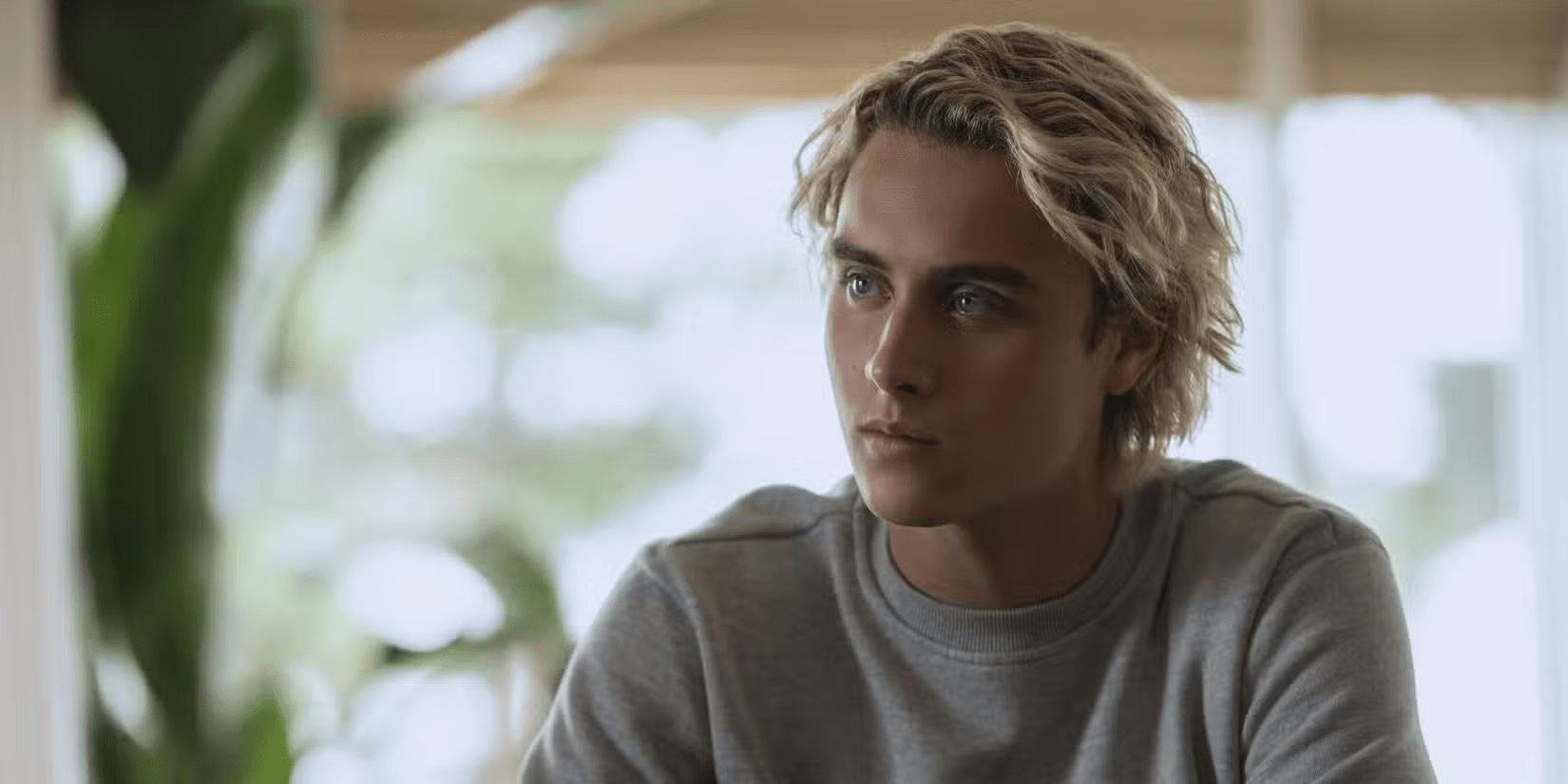 Um jovem sentado em ambiente interno, olhando com expressão séria para o lado. Ele tem cabelo loiro ondulado, um pouco comprido, e veste uma blusa de moletom cinza. Ao fundo, o cenário é desfocado, com luz natural entrando por grandes janelas e plantas visíveis, criando um clima suave e iluminado.