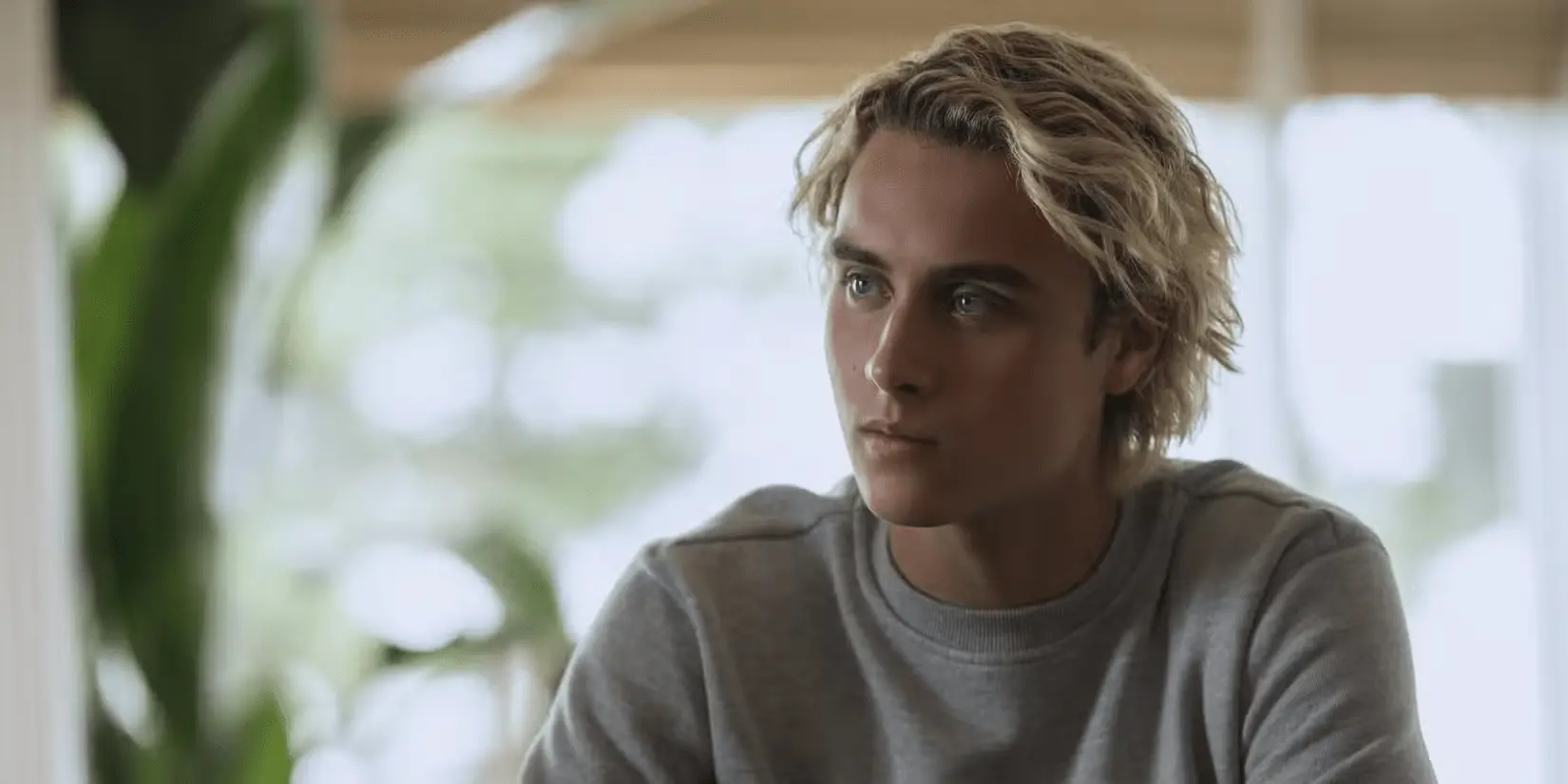 Um jovem sentado em ambiente interno, olhando com expressão séria para o lado. Ele tem cabelo loiro ondulado, um pouco comprido, e veste uma blusa de moletom cinza. Ao fundo, o cenário é desfocado, com luz natural entrando por grandes janelas e plantas visíveis, criando um clima suave e iluminado.