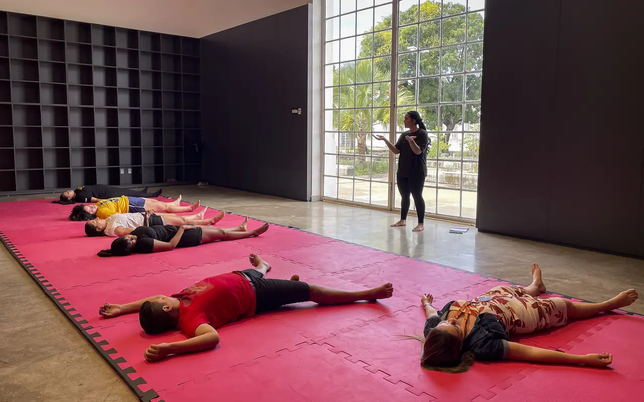 Pessoas deitadas em colchonetes vermelhos no chão de um espaço de Pinacoteca do Ceará, em uma atividade de relaxamento. Uma mulher, em pé, orienta o grupo próximo a uma grande janela que mostra a paisagem externa.