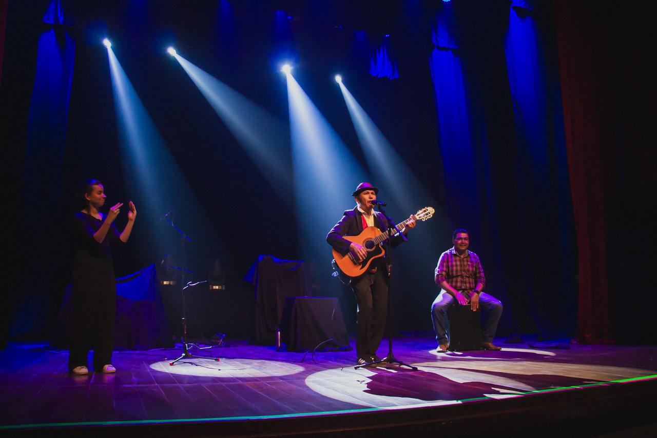 Três pessoas em um palco escuro de um teatro, iluminados por focos de luz azul e branca. Um artista ao centro canta tocando um violão, usando um chapéu. À esquerda, uma mulher faz intepretação de Libas, e à direita, um homem sentado faz a percussão.