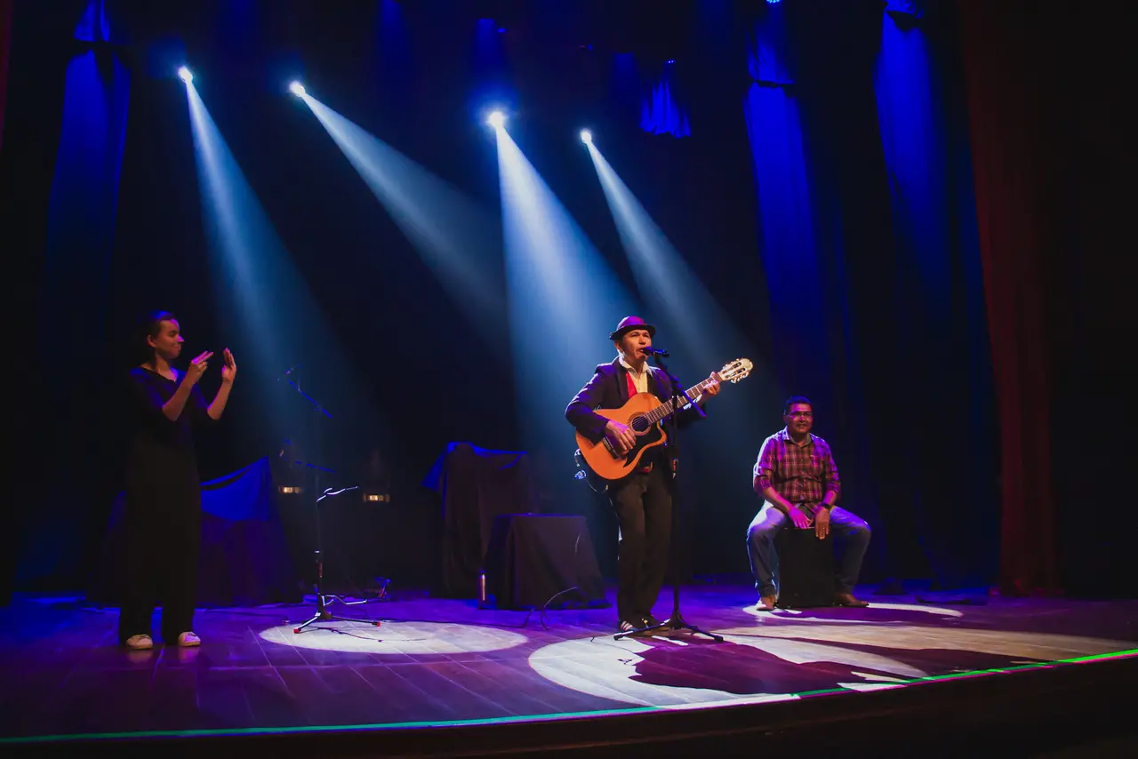 Três pessoas em um palco escuro de um teatro, iluminados por focos de luz azul e branca. Um artista ao centro canta tocando um violão, usando um chapéu. À esquerda, uma mulher faz intepretação de Libas, e à direita, um homem sentado faz a percussão.