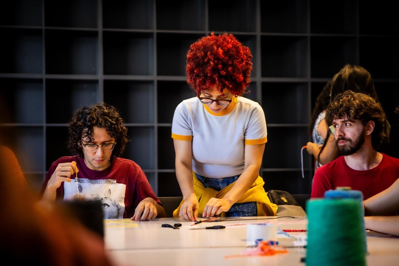 Três pessoas em uma mesa em uma oficina de artesanato. Uma mulher com cabelo crespo e ruivo está inclinada, trabalhando com linhas. Dois homens, um à esquerda e outro à direita, também participam do trabalho manual. Ao fundo, um móvel preto e vazio com espaços vazados.