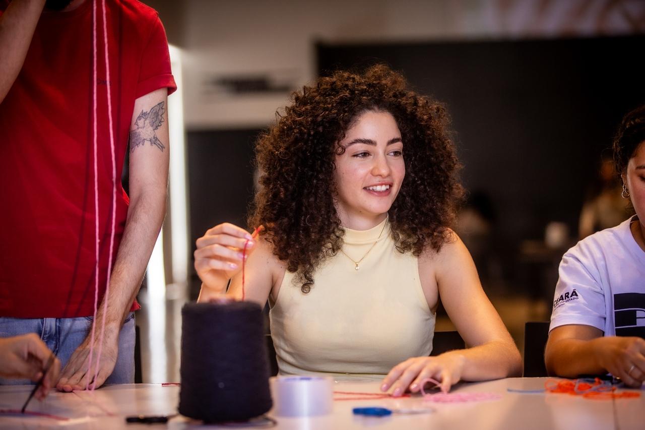 Mulher jovem de cabelo castanho encaracolado e blusa clara sentada em uma mesa, sorrindo, enquanto trabalha com linhas coloridas em uma atividade de artesanato. Na frente dela, há um grande rolo de linha escura e outros materiais. Um homem de camisa vermelha é parcialmente visível à esquerda.