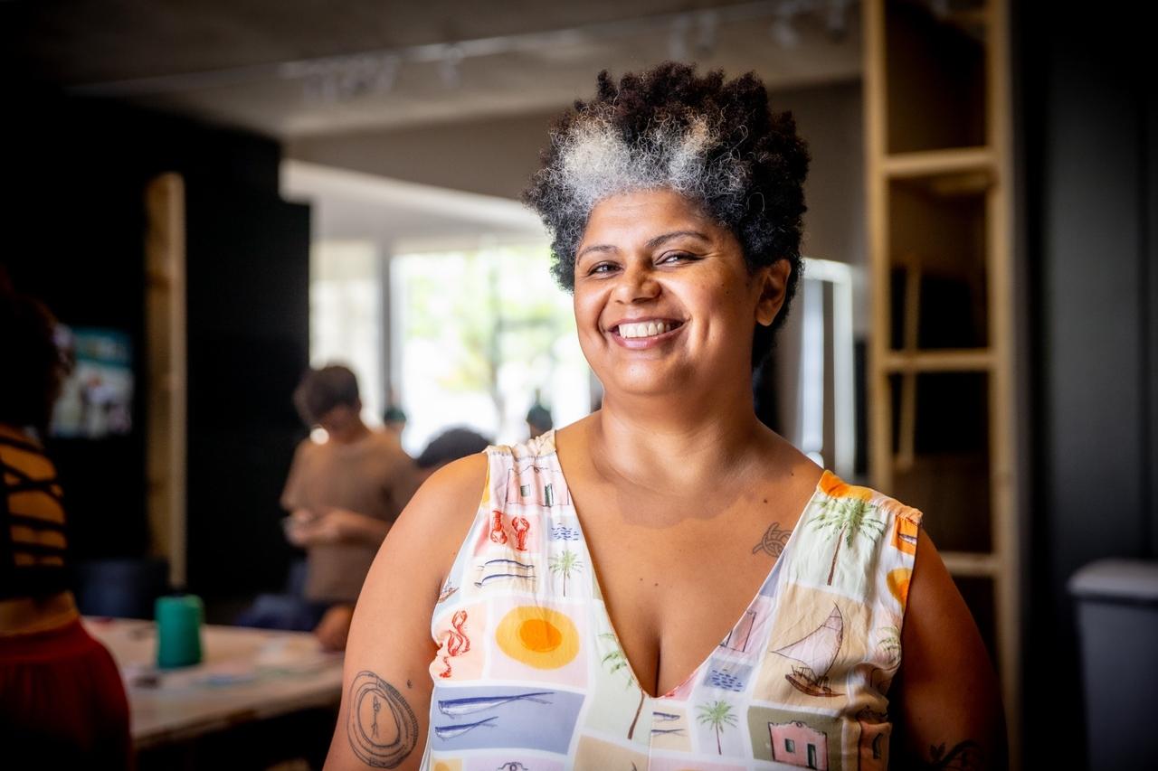 Mulher de cabelo crespo curto com mechas grisalhas frontais sorri e olha diretamente para a câmera. Ela usa uma blusa com estampa colorida de paisagens e tem tatuagens. O fundo está desfocado, mostrando um ambiente interno.