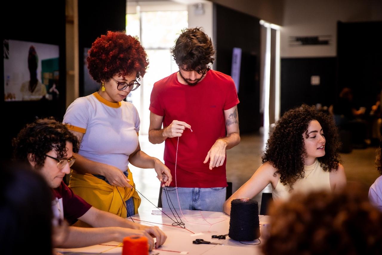Quatro pessoas em uma mesa de artesanato, manuseando linhas coloridas. A mulher de cabelo crespo ruivo e óculos e o homem de camisa vermelha estão de pé, curvados sobre o trabalho. Um homem de óculos e uma mulher de cabelo cacheado estão sentados, concentrados na atividade manual. Há um rolo grande de linha escura na mesa.