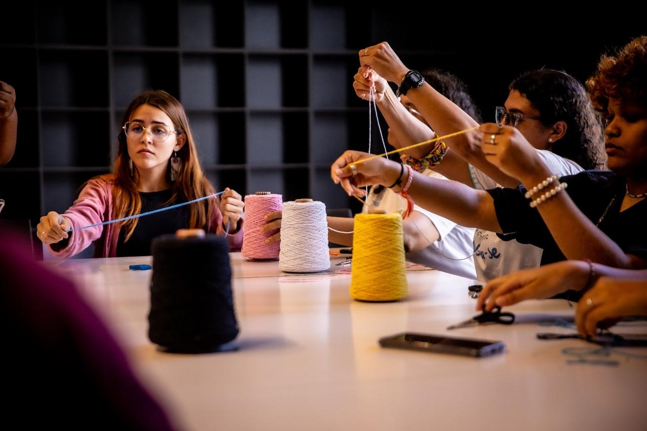 Grupo de pessoas em uma mesa de artesanato, manuseando linhas coloridas que estendem no ar. A mesa tem grandes rolos de linha preta, rosa, branca e amarela. Uma mulher de cabelo castanho e óculos, com um casaco rosa, está visível à esquerda, segurando uma linha azul clara.