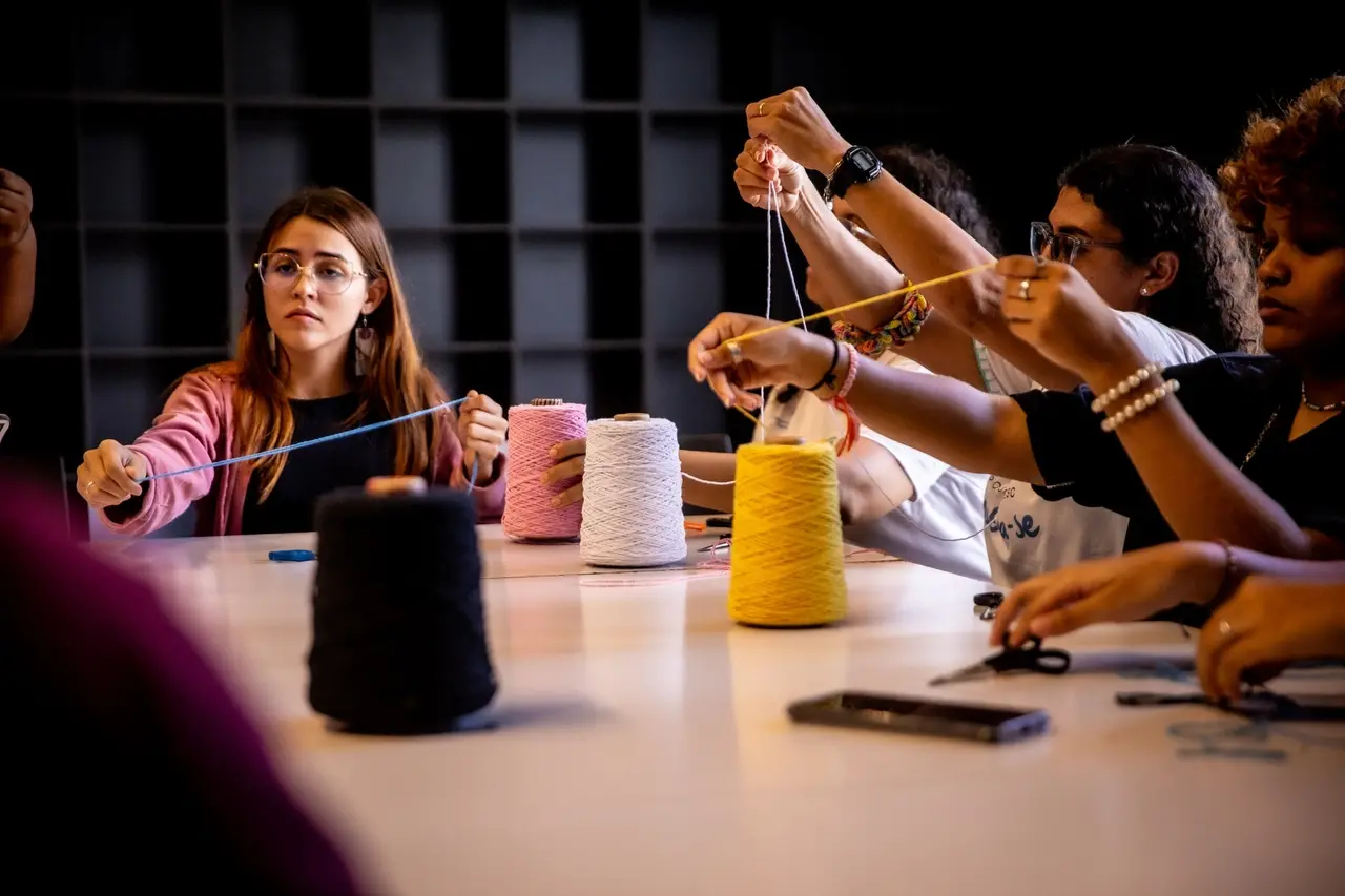 Grupo de pessoas em uma mesa de artesanato, manuseando linhas coloridas que estendem no ar. A mesa tem grandes rolos de linha preta, rosa, branca e amarela. Uma mulher de cabelo castanho e óculos, com um casaco rosa, está visível à esquerda, segurando uma linha azul clara.