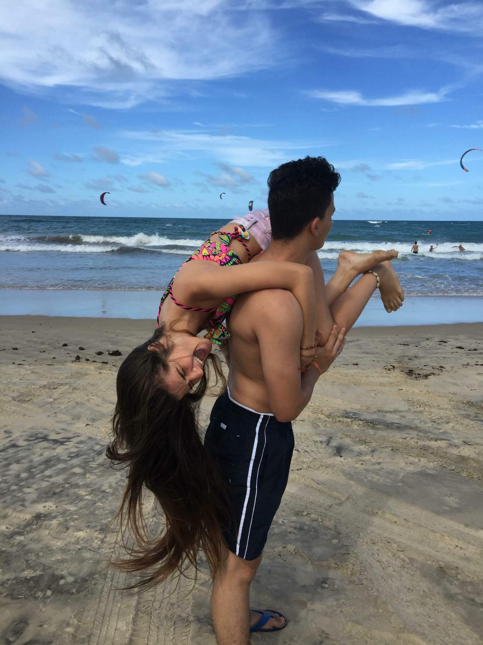 Casal de adolescentes na praia. O menino segura a menina de ponta cabeça em um momento descontraído. Ele usa uma bermuda preta e ela um short rosa com a parte de cima do biquini colorida.