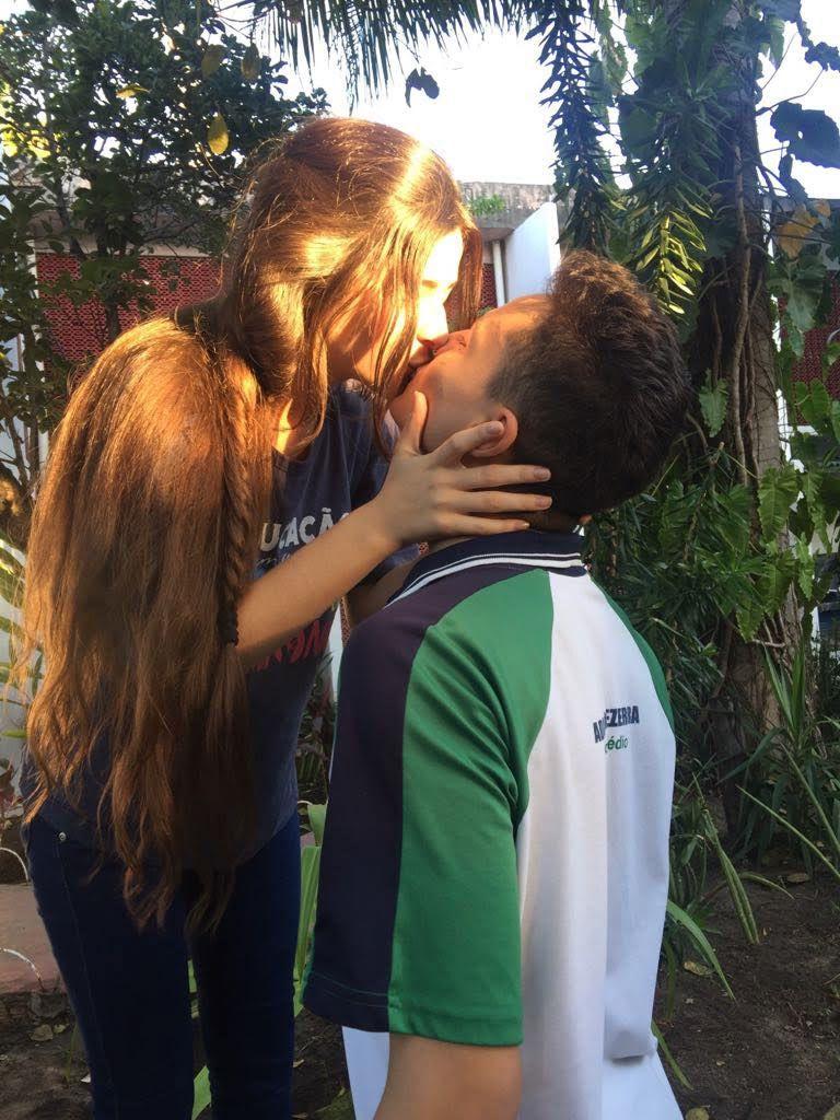 Casal de adolescentes brancos se beijando com farda de colégio nas cores azul, verde e branco. A menina tem cabelos castanhos claros e o rapaz tem cabelos pretos. Ao fundo, há plantas verdes.