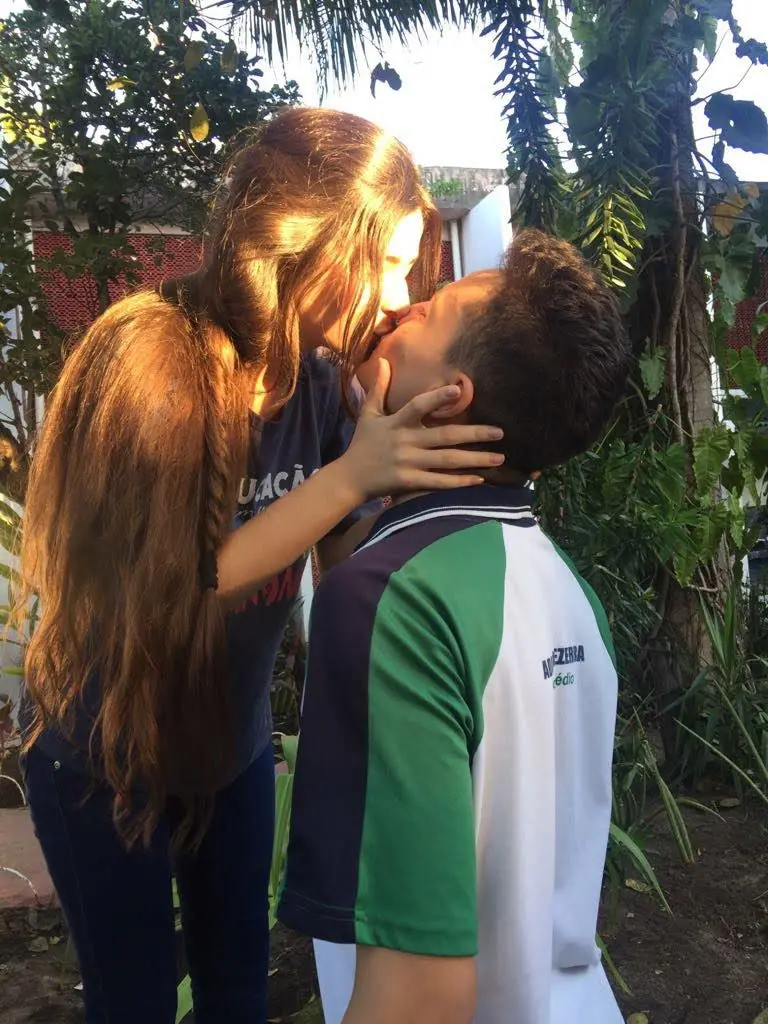 Casal de adolescentes brancos se beijando com farda de colégio nas cores azul, verde e branco. A menina tem cabelos castanhos claros e o rapaz tem cabelos pretos. Ao fundo, há plantas verdes.