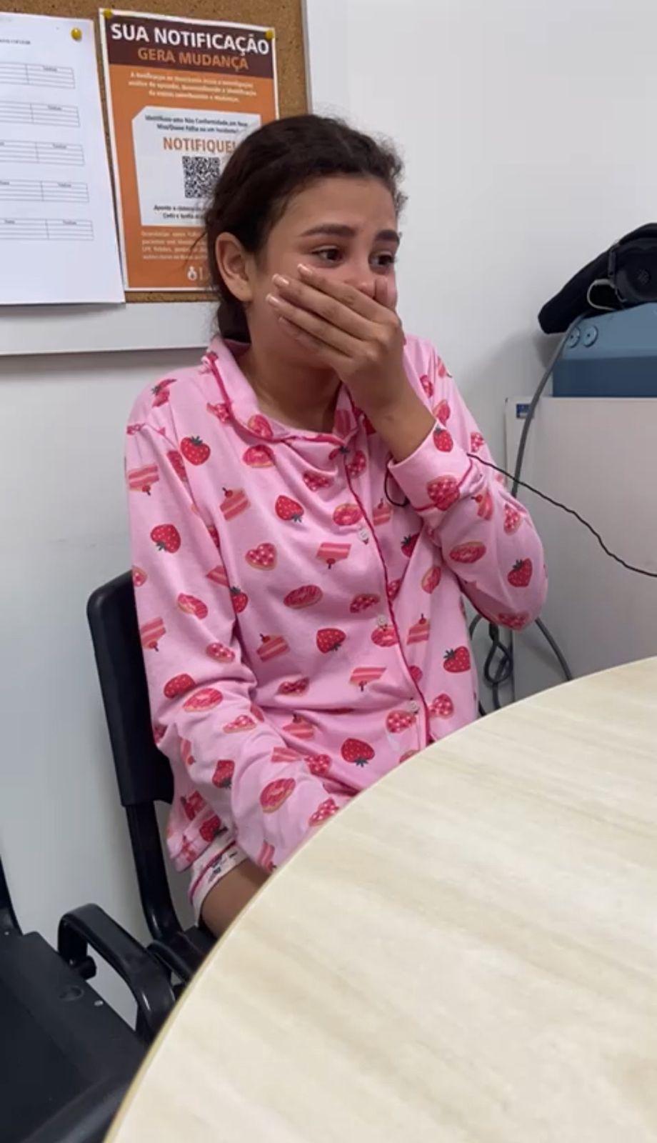 Na imagem, uma jovem, possivelmente Laís Vitória, sentada em uma cadeira preta em uma sala de escritório, vestindo um pijama rosa estampado com morangos e pedaços de bolo. Ela cobre a boca com a mão em uma expressão de surpresa e emoção, com lágrimas nos olhos, sugerindo que acabou de receber uma notícia impactante. Um mural ou quadro de avisos com papéis e um cartaz que diz 