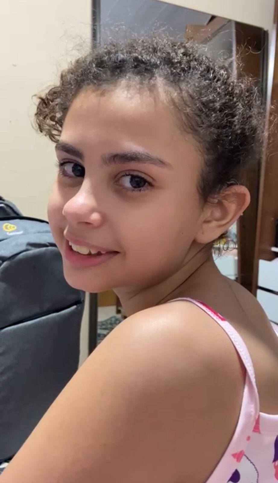 Na imagem, close-up de uma jovem com cabelo castanho cacheado, preso ou puxado para trás. Ela está usando uma alça de regata rosa com um padrão. A jovem sorri levemente e olha por cima do ombro, diretamente para a câmera. O fundo está ligeiramente desfocado, mas é possível ver uma mochila ou objeto escuro no canto esquerdo.