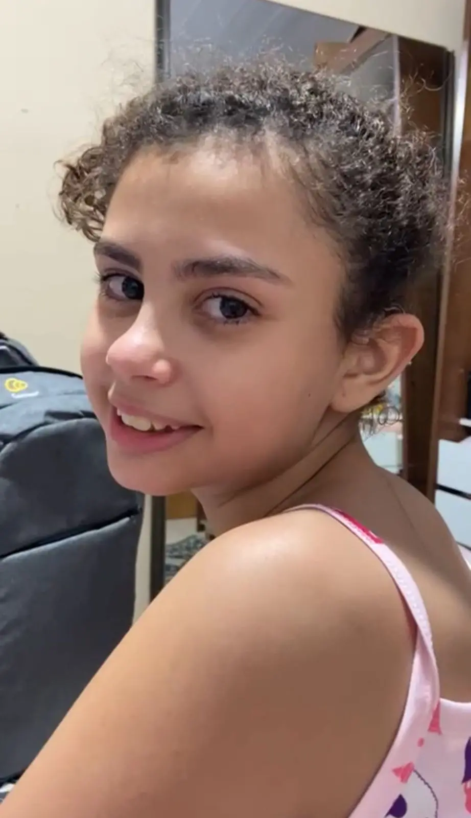 Na imagem, close-up de uma jovem com cabelo castanho cacheado, preso ou puxado para trás. Ela está usando uma alça de regata rosa com um padrão. A jovem sorri levemente e olha por cima do ombro, diretamente para a câmera. O fundo está ligeiramente desfocado, mas é possível ver uma mochila ou objeto escuro no canto esquerdo.