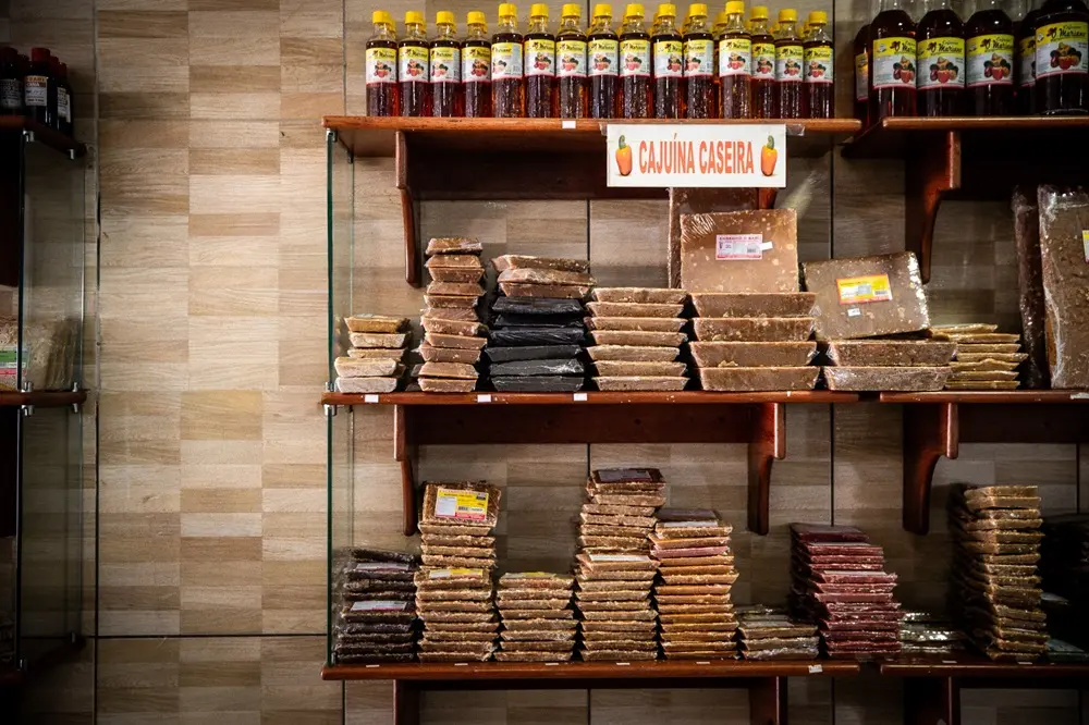 Na imagem, uma prateleira de loja bem abastecida, contra uma parede de azulejos claros em faixas, exibe pilhas de rapadura em diferentes tonalidades, desde marrom-claro a marrom-escuro e roxo. Garrafas de uma bebida chamada 