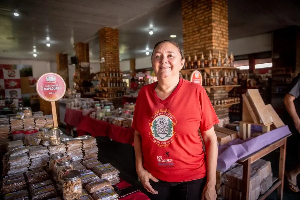 Na imagem, uma mulher sorridente em uma camiseta vermelha com um logotipo circular no peito está em pé em uma loja de produtos artesanais. Ela está cercada por pilhas de rapadura embalada e outros produtos em prateleiras e mesas, que se estendem por um amplo espaço comercial com pilares de tijolos à vista.