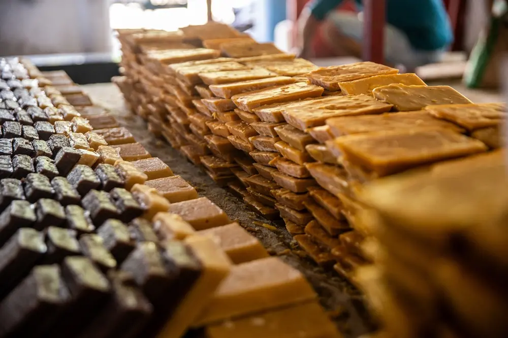 Na imagem, uma foto de perto, com baixa profundidade de campo, mostra fileiras e pilhas de rapadura. À esquerda, as peças são de uma cor marrom muito escura, e se transformam em tons mais claros, de âmbar e dourado, à medida que se movem para a direita e para o primeiro plano. Uma pessoa está fora de foco no fundo.