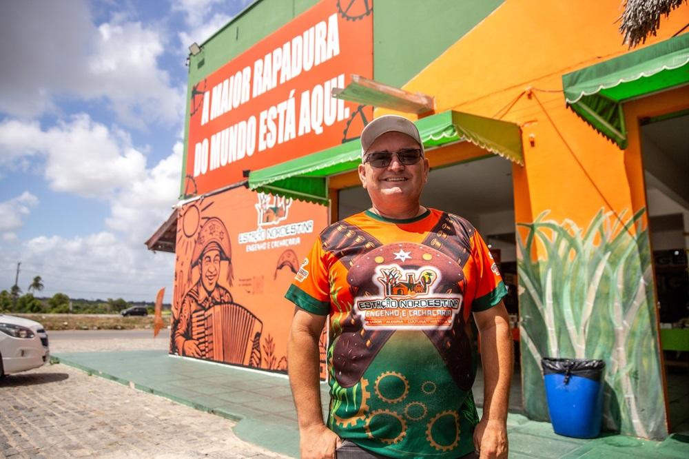 Na imagem, um homem de meia-idade, sorridente, usando óculos escuros e um boné, está em pé em frente a um edifício de cores vivas. Sua camiseta laranja e verde tem um design gráfico ousado. O edifício tem um mural de um sanfoneiro e um letreiro grande que diz, em parte,