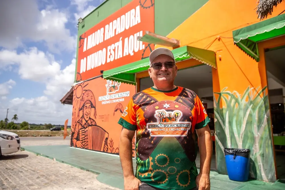 Na imagem, um homem de meia-idade, sorridente, usando óculos escuros e um boné, está em pé em frente a um edifício de cores vivas. Sua camiseta laranja e verde tem um design gráfico ousado. O edifício tem um mural de um sanfoneiro e um letreiro grande que diz, em parte, 