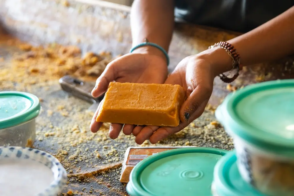Na imagem, um close-up de duas mãos segurando um pedaço retangular de rapadura de cor caramelo dourado brilhante, possivelmente saborizada. As mãos usam pulseiras e estão sobre uma superfície coberta de migalhas do doce. Recipientes de plástico com tampas verde-água e outros ingredientes estão no primeiro plano.