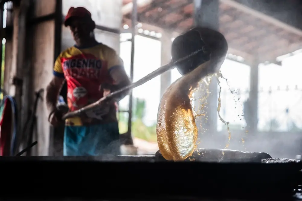Na imagem, uma foto com foco em um plano próximo registra o processo de fabricação de rapadura em um engenho. Um líquido espesso e brilhante, de cor caramelo vibrante, escorre de uma grande concha preta, formando um filete curvo e viscoso que cai sobre uma superfície escura. Gotas de calda espirram ao redor. Em segundo plano e desfocado, um homem com um boné vermelho e uma camiseta colorida que diz 
