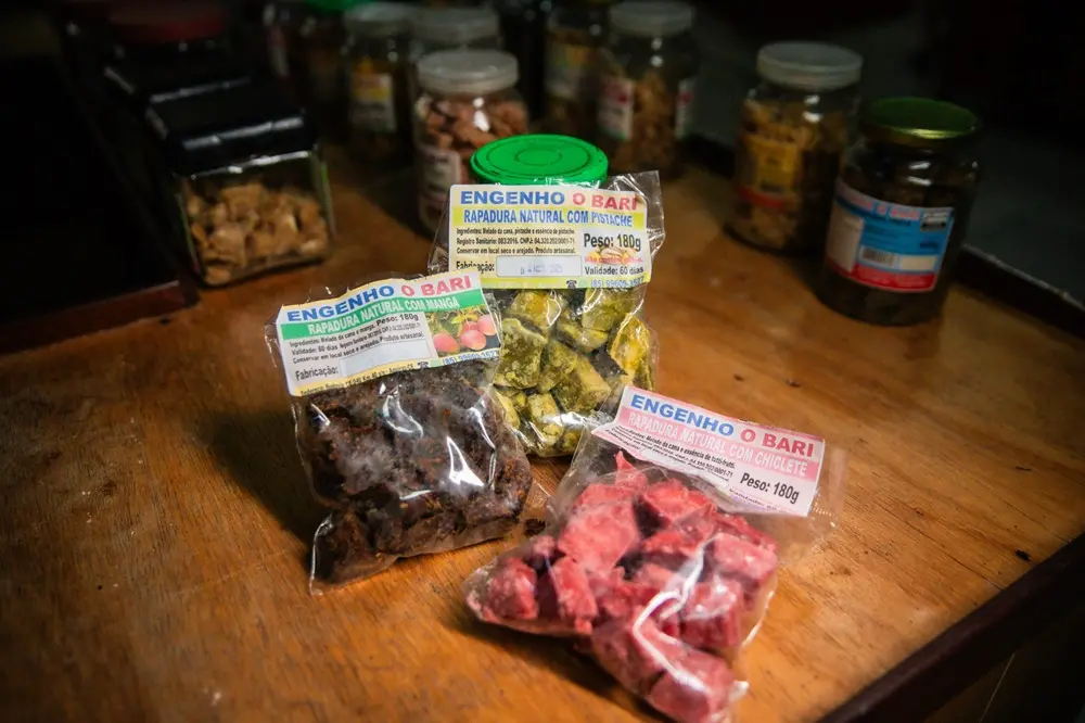 Na imagem, uma foto em close-up, com iluminação focalizada e profundidade de campo rasa, mostra três embalagens plásticas transparentes de rapadura artesanal em pedaços sobre uma mesa de madeira. Cada embalagem contém um sabor diferente: Embalagem superior (Verde): Rapadura com pistache, de cor verde; embalagem esquerda (Marrom Escuro): Rapadura com manga, de cor marrom escura (quase preta); embalagem inferior direita (Vermelha): Rapadura com chiclete, de cor rosa/vermelha intensa. Todas as embalagens têm um rótulo que identifica o produto como 