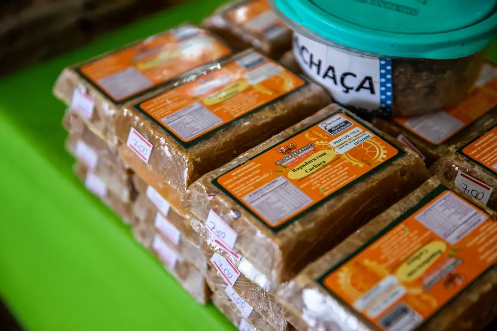 Na imagem, uma foto em close-up, com ângulo inclinado, mostra pilhas de rapadura retangular e embalada individualmente em plástico, sobre uma superfície verde neon. A rapadura tem uma cor caramelo-dourada translúcida e possui um rótulo laranja vibrante. O rótulo de uma das barras é legível e diz 