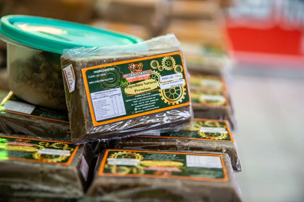 Na imagem, uma foto em close-up, com profundidade de campo rasa, exibe várias barras retangulares de rapadura escura empilhadas, todas embaladas individualmente em plástico. Em primeiro plano, uma das barras está inclinada, exibindo claramente o rótulo principal. O rótulo tem uma moldura laranja e um fundo escuro com desenhos de engrenagens (sugerindo 